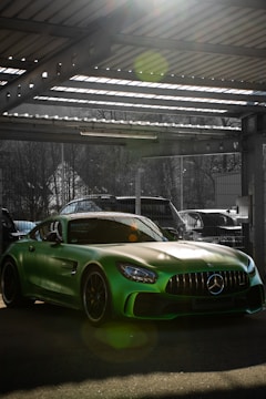 A sleek green sports car is parked in a sunlit garage. The car's glossy paint reflects the light, creating vibrant lens flares. Metal beams and a corrugated roof are visible above, with some trees and a wire fence in the background.