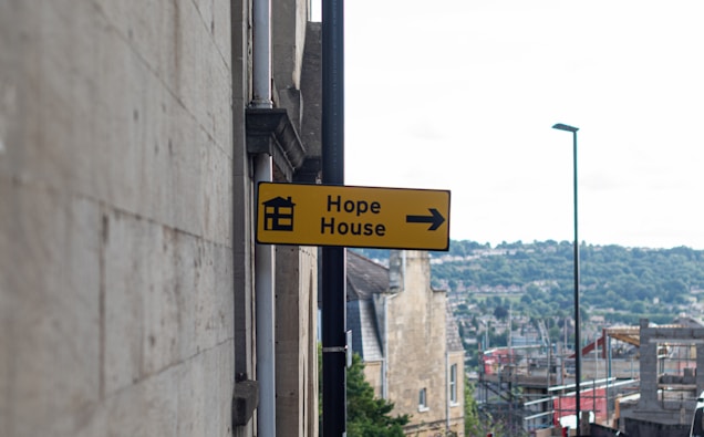A yellow sign with a black border is attached to the side of a stone building. It features an icon of a house and the words 'Hope House' alongside an arrow pointing to the right. The background shows a cityscape with hills and a cloudy sky.