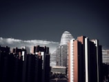 A skyline featuring tall modern buildings, including a distinctive cylindrical structure with horizontal lines. The foreground includes high-rise buildings predominantly in dark colors, contrasted by a clear blue sky with a strip of white clouds.
