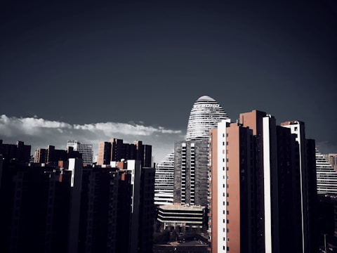 A skyline featuring tall modern buildings, including a distinctive cylindrical structure with horizontal lines. The foreground includes high-rise buildings predominantly in dark colors, contrasted by a clear blue sky with a strip of white clouds.