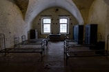 A dimly lit room with arched ceilings and a rustic, aged appearance. The room contains several metal-framed beds arranged parallel to each other, with a couple of dark storage cabinets along the walls. The stone walls have a weathered look, and a single window lets in limited natural light.