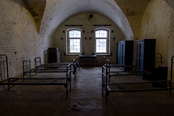 A dimly lit room with arched ceilings and a rustic, aged appearance. The room contains several metal-framed beds arranged parallel to each other, with a couple of dark storage cabinets along the walls. The stone walls have a weathered look, and a single window lets in limited natural light.