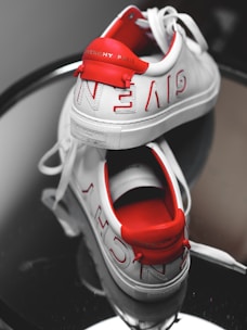 A pair of stylish, white Givenchy sneakers placed on a reflective surface. The sneakers have red accents and show the Givenchy Paris branding prominently on the back heel and along the sides. One shoe is positioned upright while the other lies on its side, revealing the insole.