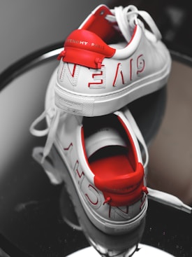 A pair of stylish, white Givenchy sneakers placed on a reflective surface. The sneakers have red accents and show the Givenchy Paris branding prominently on the back heel and along the sides. One shoe is positioned upright while the other lies on its side, revealing the insole.