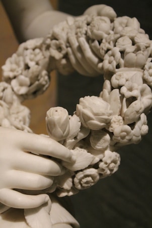 A detailed marble sculpture features intricately carved hands holding a garland of various flowers and leaves. The craftsmanship highlights delicate textures and floral details.