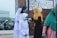 A group of women and girls wearing colorful hijabs and masks are standing outside near a gate. One woman in a yellow hijab is using a handheld thermometer to check the temperature of a girl in a white hijab and outfit. Several motorcycles are parked in the background.