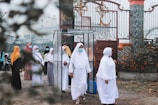 A group of women wearing hijabs and face masks gather near a decorative metal gate. They seem to be walking through a transparent booth situated at the entrance of a walled compound. The area is adorned with greenery and flowering plants.