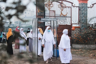 A group of women wearing hijabs and face masks gather near a decorative metal gate. They seem to be walking through a transparent booth situated at the entrance of a walled compound. The area is adorned with greenery and flowering plants.