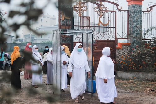 A group of women wearing hijabs and face masks gather near a decorative metal gate. They seem to be walking through a transparent booth situated at the entrance of a walled compound. The area is adorned with greenery and flowering plants.