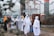 A group of women wearing hijabs and face masks gather near a decorative metal gate. They seem to be walking through a transparent booth situated at the entrance of a walled compound. The area is adorned with greenery and flowering plants.