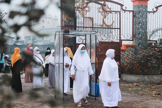 A group of women wearing hijabs and face masks gather near a decorative metal gate. They seem to be walking through a transparent booth situated at the entrance of a walled compound. The area is adorned with greenery and flowering plants.
