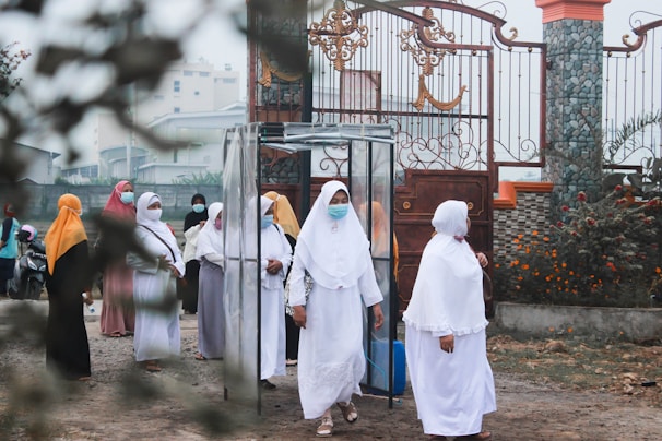 A group of women wearing hijabs and face masks gather near a decorative metal gate. They seem to be walking through a transparent booth situated at the entrance of a walled compound. The area is adorned with greenery and flowering plants.