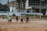 A group of people are playing beach volleyball on a sandy court. They are engaged in a game, with a ball in mid-air above the net. The background features a partially constructed or abandoned building, with some palm trees lining the area. The setting appears to be a seaside or coastal location, and the weather seems mild.