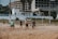 A group of people are playing beach volleyball on a sandy court. They are engaged in a game, with a ball in mid-air above the net. The background features a partially constructed or abandoned building, with some palm trees lining the area. The setting appears to be a seaside or coastal location, and the weather seems mild.