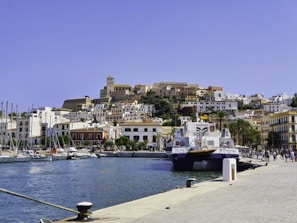 A picturesque harbor with boats docked along clear blue waters. In the background, a charming hillside town with white buildings and a prominent historical structure at the top. Palm trees line the waterfront, adding a tropical feel. People stroll along the quay, enjoying the sunny weather.
