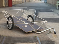 A minimalist, two-wheeled cart with a sleek metal frame and a flat wooden surface. It is situated on an urban street, with buildings and industrial elements in the background.