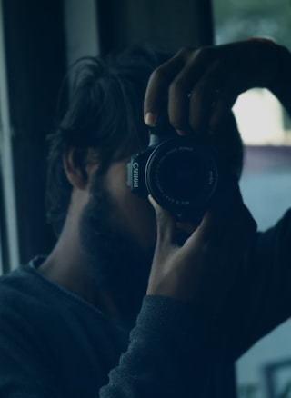 A person holding a camera close to their face, taking a photo in a dimly lit room. The focus is on the camera, which is a Canon, while the person is partially obscured and in shadow. There is a window in the background letting in natural light.