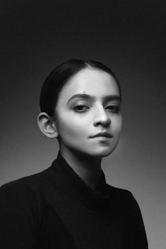 A black and white portrait of a person with a calm and composed expression dressed in a dark turtleneck against a softly lit background. The lighting emphasizes the contours of their face, creating a dramatic and classic look.