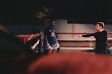 A man wearing boxing gloves and a blue shirt stands inside a boxing ring. Another man, standing outside the ring, appears to be giving instructions or coaching him. The environment is dimly lit, suggesting an indoor gym or training facility.