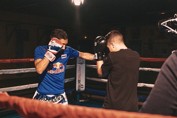 Two men are engaged in a boxing match inside a ring. One is wearing a blue outfit with a logo on the shirt, and the other is dressed in black. Both have raised gloves, preparing to spar.