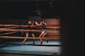 Two men are engaged in a sparring session inside a boxing ring, with one wearing black shorts and gloves and the other in white shorts and blue top. The ring is dimly lit, creating a focused and intense atmosphere.