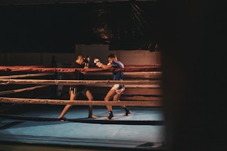 Two men are engaged in a sparring session inside a boxing ring, with one wearing black shorts and gloves and the other in white shorts and blue top. The ring is dimly lit, creating a focused and intense atmosphere.