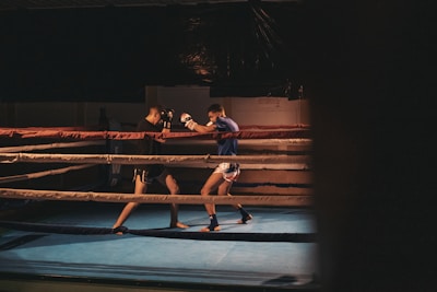 Two men are engaged in a sparring session inside a boxing ring, with one wearing black shorts and gloves and the other in white shorts and blue top. The ring is dimly lit, creating a focused and intense atmosphere.