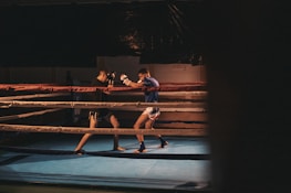 Two men are engaged in a sparring session inside a boxing ring, with one wearing black shorts and gloves and the other in white shorts and blue top. The ring is dimly lit, creating a focused and intense atmosphere.