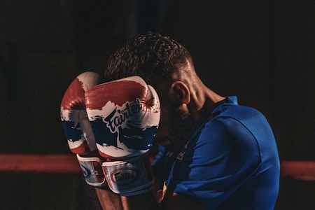 A person wearing boxing gloves is in a defensive stance, with their arms raised to cover their face. The gloves are brightly colored with a red, white, and blue design, and the word 'Fairtex' is visible on them. The scene is dimly lit, with the person wearing a blue athletic shirt.