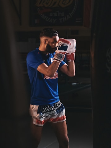 A man in a blue athletic shirt and sporty shorts is standing in a fighting pose, wearing boxing gloves inside a dimly lit gym setting. The backdrop suggests a focus on martial arts, with a Muay Thai related banner partially visible.