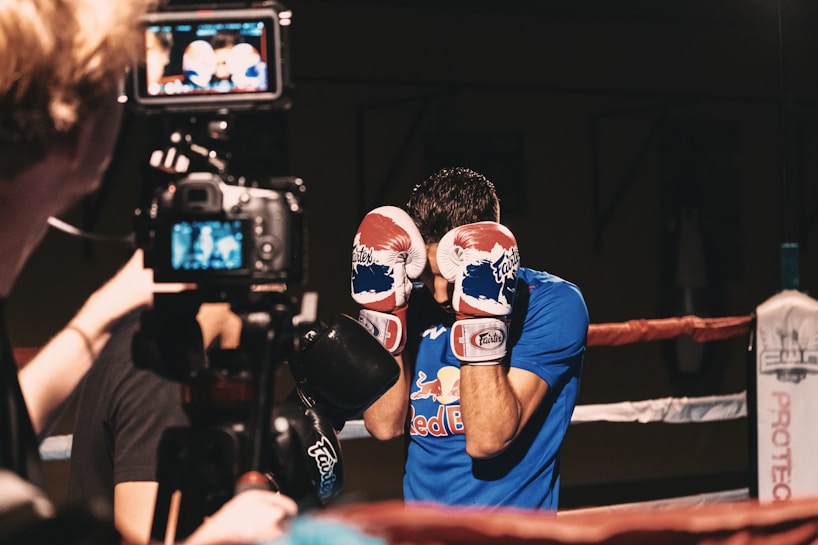 A man wearing boxing gloves stands inside a boxing ring, bringing his gloves up to protect his face. The scene is captured by a camera operated by another person nearby. He wears a blue shirt with a Red Bull logo. The boxing ring is enclosed by red and white ropes.