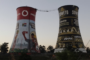 Two large cooling towers are visible, each decorated with colorful murals and advertisements. One tower features vibrant artwork including a South African flag, people taking selfies, and a red section with a prominent brand logo. The other tower is adorned with black and gold imagery, displaying words associated with Soweto, alongside various cultural and local symbols.