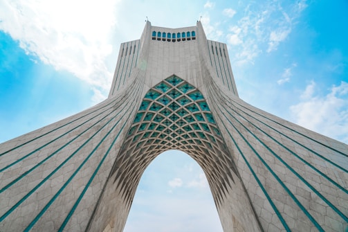 A large architectural structure with an archway, featuring geometric patterns and vertical lines. The structure is made of white materials with teal accents and is set against a partly cloudy blue sky.