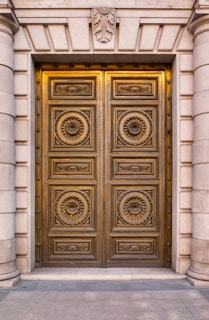 A grand, ornate double door made of metal with intricate circular patterns and decorative motifs featuring floral designs. The door is set in a stone facade with large, vertical columns flanking each side. Above the door is an emblem with leaves, adding to the classical architectural style.