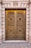 A grand, ornate double door made of metal with intricate circular patterns and decorative motifs featuring floral designs. The door is set in a stone facade with large, vertical columns flanking each side. Above the door is an emblem with leaves, adding to the classical architectural style.