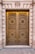 A grand, ornate double door made of metal with intricate circular patterns and decorative motifs featuring floral designs. The door is set in a stone facade with large, vertical columns flanking each side. Above the door is an emblem with leaves, adding to the classical architectural style.