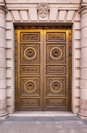 A grand, ornate double door made of metal with intricate circular patterns and decorative motifs featuring floral designs. The door is set in a stone facade with large, vertical columns flanking each side. Above the door is an emblem with leaves, adding to the classical architectural style.