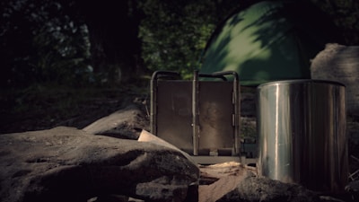 A green tent is set up in a wooded area with a metal camping stove and a stainless steel container visible in the foreground. Large rocks and a tree trunk are scattered around the campsite, with dense foliage in the background. The scene is dimly lit, suggesting early morning or late evening.
