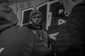 A moody black-and-white photo of a model wearing an oversized black hoodie, standing against a graffiti-covered urban wall at night.