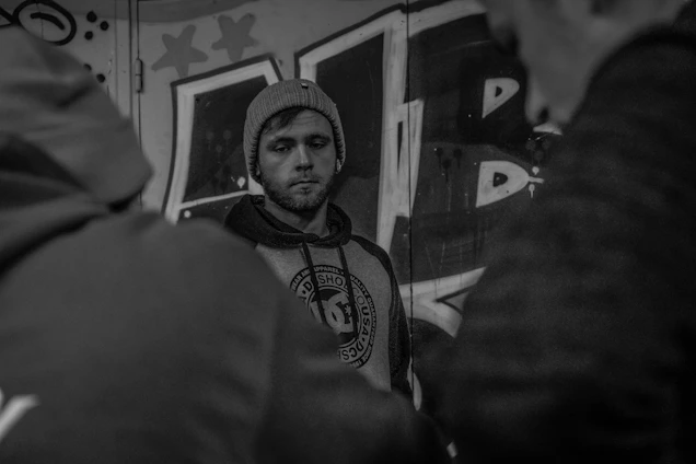 A moody black-and-white photo of a model wearing an oversized black hoodie, standing against a graffiti-covered urban wall at night.