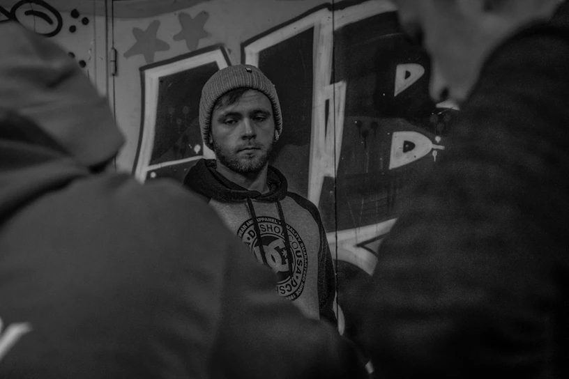 A moody black-and-white shot of a model wearing an oversized charcoal hoodie with silver accents, standing against a graffiti-covered wall at night.