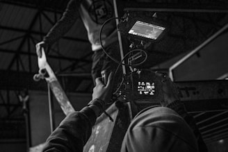 A black and white scene of a person holding a video camera, focusing on a skateboarder performing a trick on a ramp. The setting appears to be indoors, possibly a skate park with structural beams visible in the background. The skateboarder is captured mid-action, adding dynamic movement to the composition.