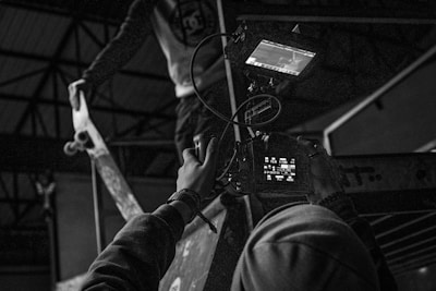 A black and white scene of a person holding a video camera, focusing on a skateboarder performing a trick on a ramp. The setting appears to be indoors, possibly a skate park with structural beams visible in the background. The skateboarder is captured mid-action, adding dynamic movement to the composition.