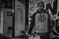 A person stands in an urban setting, dressed in a hoodie and knit cap, holding a skateboard. The background features graffiti-covered walls and a small wooden structure with a speaker on top. The scene conveys a gritty, street-style atmosphere.