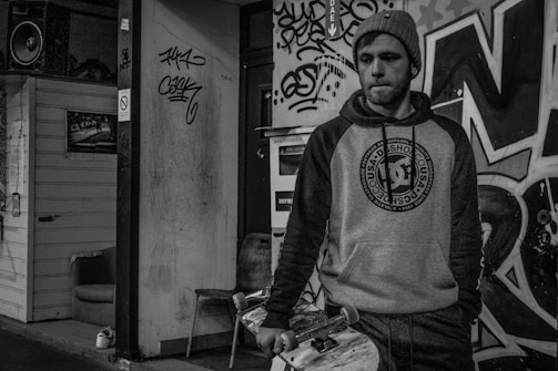 A person stands in an urban setting, dressed in a hoodie and knit cap, holding a skateboard. The background features graffiti-covered walls and a small wooden structure with a speaker on top. The scene conveys a gritty, street-style atmosphere.