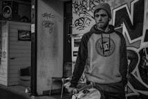 A person stands in an urban setting, dressed in a hoodie and knit cap, holding a skateboard. The background features graffiti-covered walls and a small wooden structure with a speaker on top. The scene conveys a gritty, street-style atmosphere.