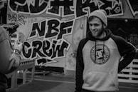 A person in a beanie and hoodie with a streetwear design is standing in front of a wall covered in graffiti art. The graffiti includes bold letters and intricate patterns. Another person is partially visible, holding a camera.
