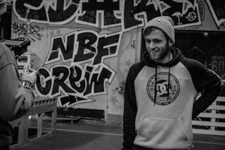 A person in a beanie and hoodie with a streetwear design is standing in front of a wall covered in graffiti art. The graffiti includes bold letters and intricate patterns. Another person is partially visible, holding a camera.