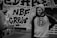 A person in a beanie and hoodie with a streetwear design is standing in front of a wall covered in graffiti art. The graffiti includes bold letters and intricate patterns. Another person is partially visible, holding a camera.