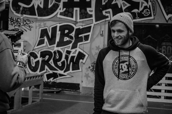 A person in a beanie and hoodie with a streetwear design is standing in front of a wall covered in graffiti art. The graffiti includes bold letters and intricate patterns. Another person is partially visible, holding a camera.